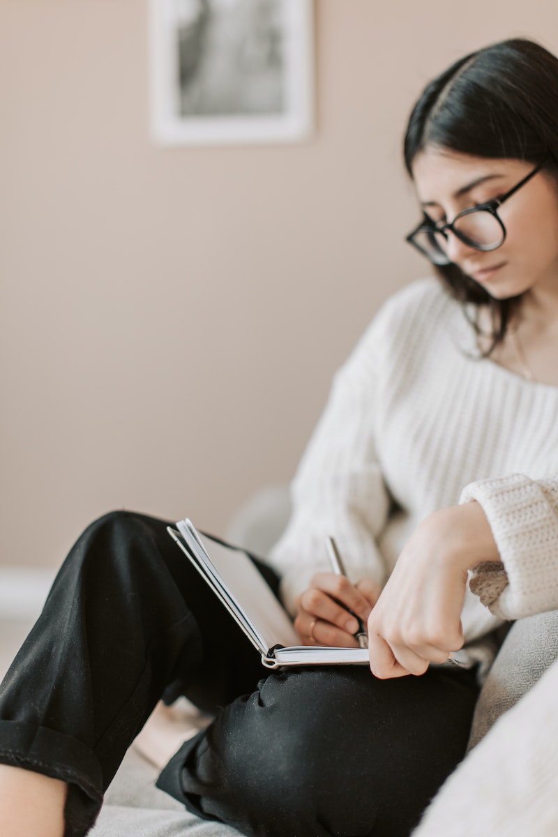 Attentive female wearing eyeglasses and casual outfit sitting barefoot with crossed legs on comfortable couch in modern flat and taking notes in notebook with pen