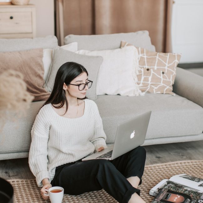 Focused female in casual wear sitting on floor carpet near sofa with cup of tea and browsing laptop while spending time at home